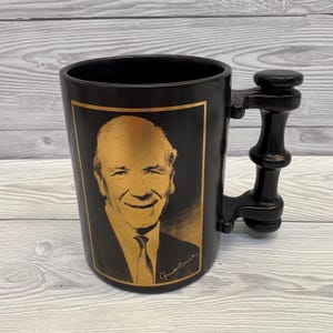 May include: Black ceramic mug with a gold-toned portrait of a smiling man in a suit and tie. The mug has a distinctive handle and a gold border around the portrait. The man's signature is at the bottom of the portrait.