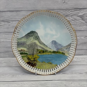 Può includere: Piatto decorativo con un paesaggio montano, un lago e alberi. Il bordo bianco presenta dettagli dorati e un motivo traforato. È esposto su un supporto argentato. La scena è dipinta in tonalità tenui.