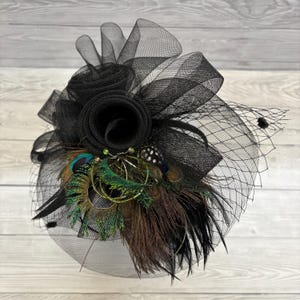 Könnte beinhalten: Ein schwarzer Fascinator mit einem Netzschleier, einer großen Stoffrose und einer Schleife. Der Kopfschmuck ist mit Pfauenfedern in Grün-, Braun- und Blautönen verziert. Die Basis ist hellgrau.