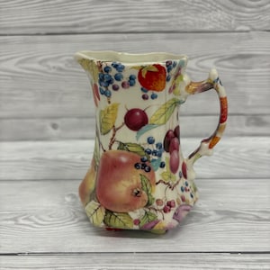 Puede incluir: Jarra de cerámica decorativa con diseño de frutas. La jarra presenta un fondo crema con ilustraciones de manzanas, cerezas, ciruelas y arándanos. El asa es multicolor y la jarra tiene una forma única.
