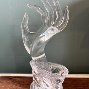 Puede incluir: Una escultura de mano de cristal transparente con los dedos extendidos. La mano emerge de una base decorativa y texturizada. La escultura está diseñada para sostener joyas o artículos de exhibición. El cristal transparente refleja la luz.