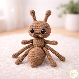 May include: A crocheted brown ant with large black eyes, antennae, and six legs. The amigurumi ant is made of yarn and has a rounded head and body. The handmade toy is sitting on a white surface.