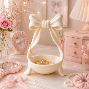May include: A cream-coloured jewellery stand featuring a large bow at the top and a circular base. The stand holds a gold necklace with a pink heart pendant and a gold hoop earring with a small pink bow and pearl charm. Several gold rings sit inside.