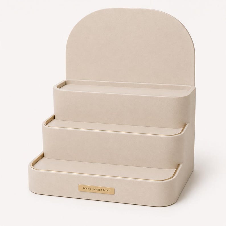 May include: A beige, three-tiered display stand with an arched top. The stand has a rectangular base and rounded edges. A small rectangular label at the base reads "SCENT YOUR STORY".