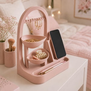 Op de afbeelding: Een roze sieraden- en accessoire-organizer met een boogvormig ontwerp. Het heeft een bovenste balk om sieraden op te hangen, twee ovale bakjes, een telefoonhouder en een bakje voor pennen. Gouden sieraden en haaraccessoires worden getoond.
