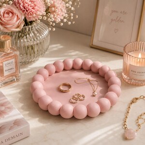 Op de afbeelding: Een rond roze decoratief dienblad met een rand van uniforme houten bollen. Het dienblad bevat een goudkleurige ring, een fijne gouden ketting met een kleine parelhanger en twee kleine gouden oorringen, allemaal gerangschikt op een vlak oppervlak.