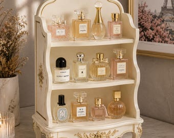 May include: A cream-colored, three-tiered wooden display shelf with gold-toned floral carvings and curved side panels. The shelf holds various glass perfume bottles of different shapes and sizes, arranged across three open storage levels.