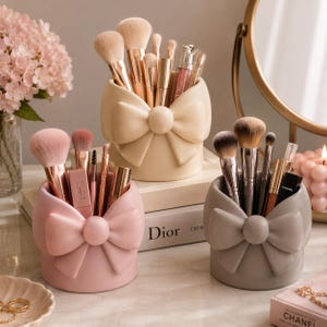 May include: Three cylindrical makeup brush holders in cream, light pink, and grey, each featuring a prominent sculpted bow on the front. The holders contain various makeup brushes, lip products, and a tube with Dior branding visible on a book.