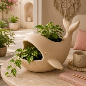 May include: A matte beige ceramic whale-shaped planter featuring a hollow opening for plants, a raised tail, and a side fin. The planter holds a green and white variegated pothos plant with trailing vines extending from the whale's mouth.