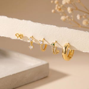 Könnte beinhalten: Ein Set aus fünf goldfarbenen Ohrringen, bestehend aus einem kleinen Schleifenstecker, einem hängenden Kristallanhänger, einem mit Kristallen besetzten Creolen-Ohrring, einem schlangenförmigen Ohrring mit grünem Auge und einer schlichten, polierten Creole.