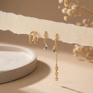 May include: Gold-toned earrings featuring a celestial theme. The set includes a pair of huggie hoop earrings, and two pairs of dangle earrings with crescent moon and star designs. The earrings are adorned with clear and green gemstones.