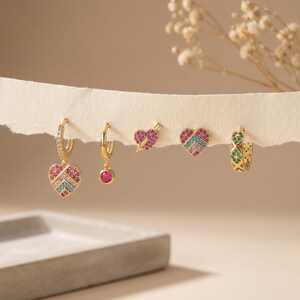 Könnte beinhalten: Ein Set aus fünf goldfarbenen Ohrringen mit verschiedenen Designs: ein herzförmiger Anhänger mit rosa und blauen Steinen, ein kleiner Creolen-Ohrring mit einem rosa Stein, ein von einem Pfeil durchbohrtes Herz, ein Herz mit rosa und blauen Steinen und ein grüner Creolen-Ohrring.