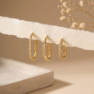 May include: Three gold-coloured hoop earrings. Two are oval-shaped and encrusted with small, clear stones. The third earring has a rectangular design with a row of clear stones. The earrings are displayed on a piece of torn, off-white paper.