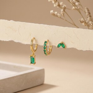 Può includere: Un set di tre orecchini dorati. Un paio presenta un pendente rettangolare con pietra verde smeraldo. Un altro paio ha una fila di pietre verde smeraldo. Il terzo paio ha due pietre a forma di marquise verde smeraldo.