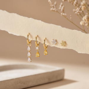 Könnte beinhalten: Ein Set aus fünf goldfarbenen Ohrringen, bestehend aus drei Creolen mit hängenden Anhängern und zwei Ohrsteckern. Die Anhänger umfassen klare und gelbe birnenförmige Steine sowie einen quadratischen Stein. Die Ohrstecker sind blumenförmig.