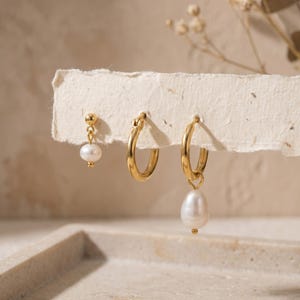 May include: Three gold earrings displayed on a textured, off-white surface. The earrings include a stud with a dangling pearl, a small hoop, and a larger hoop with a teardrop-shaped pearl pendant. The jewellery is set against a neutral background.
