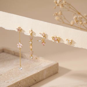 Könnte beinhalten: Ein Set aus fünf goldfarbenen Ohrringen mit zarten Blumen- und Schmetterlingsmotiven. Die Designs umfassen einen langen Kettenanhänger mit Blume, eine vertikale Sternenkette, ein kleines Blumencluster, eine Schleife und einen kleinen Schmetterling mit rosa Steinen.