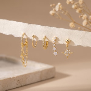 Könnte beinhalten: Ein Set aus fünf goldfarbenen Ohrringen mit verschiedenen Designs, darunter ein Creolen-Ohrring mit zwei hängenden Ketten, ein schlichter Creolen-Ohrring, ein pavé-besetzter Creolen-Ohrring, ein gebogener Stab mit abgestuften Steinen und ein Stern-Ohrring.