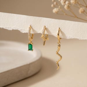 May include: Three gold-colored earrings. One earring features a green emerald-cut stone, another a lightning bolt, and the third a snake design. The earrings are displayed on a white surface with a neutral background.