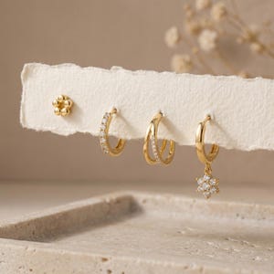 May include: A collection of gold earrings displayed on a white card. The set includes a small flower stud, a hoop earring with clear stones, a double hoop earring, and a hoop earring with a dangling flower pendant.