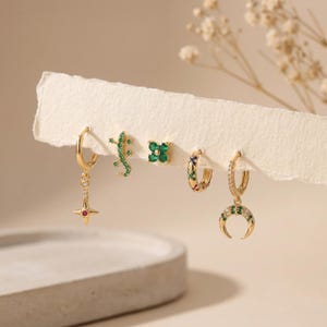 Könnte beinhalten: Ein Set aus fünf goldfarbenen Ohrringen, bestehend aus einem Sternanhänger mit rotem Mittelstein, einem grünen echsenförmigen Stecker, einem vierblättrigen Kleeblatt-Stecker mit grünen Steinen, einem kleinen Reifen mit bunten Steinen und einem Mond.