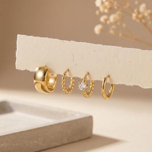 Könnte beinhalten: Ein Set aus vier goldfarbenen Creolen, die auf einem weißen Papierstreifen präsentiert werden. Das Set besteht aus einer dicken, glatten Creole, zwei perlenartigen Creolen und einer perlenartigen Creole mit einem klaren, runden Stein.