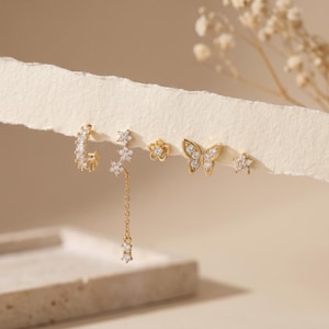 Könnte beinhalten: Ein Set aus fünf goldfarbenen Ohrringen mit klaren Steinen. Die Kollektion umfasst einen kleinen Creolen-Ohrring, eine Blumen-Cluster-Form mit einer hängenden Kette, einen Blumenstecker, einen Schmetterlingsstecker und einen kleinen Sternstecker.