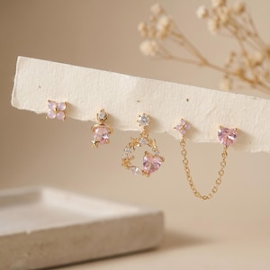 Op de afbeelding: Een verzameling delicate oorbellen in goud en roze. De set bevat oorstekers met een bloemmotief, een planeetvormige oorbel met een ster, een hartvormige oorbel en een dubbele oorbel met een hart en een bloem.