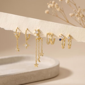 Könnte beinhalten: Ein Set aus fünf goldfarbenen Ohrringen mit Himmelsmotiven wie Sternen und einem Halbmond. Die Designs enthalten klare Zirkonia-Steine, einen kleinen blauen Stein und zarte Ketten. Die Ohrringe sind in einer Reihe angeordnet.