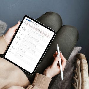 May include: A person is holding a tablet with a white stylus. The tablet displays a document with text and symbols. The person is wearing a tan sweater and dark green pants, sitting in a wicker chair.