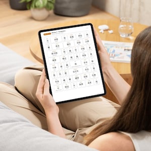 May include: A person holding a black tablet displaying a vocabulary lesson. The screen shows a grid of Chinese characters with English translations. The tablet is held in front of the person, who is sitting on a couch. The background includes a table and a plant.