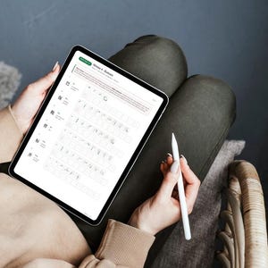 May include: A person holding a tablet with a white stylus. The tablet displays a digital document with text and symbols. The person is wearing a beige sweater and sitting on a chair with dark green pants.