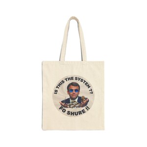 Peut inclure: Sac fourre-tout crème avec un graphisme circulaire représentant un homme portant des lunettes de soleil tenant de l'argent. Le texte autour du graphisme se lit "IS THIS THE SYSTEM?? FO SHURE !!". Le sac a un design simple avec deux poignées.