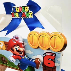 Op de afbeelding: Een witte geschenkdoos versierd met een blauw lint en een kleurrijk "Super Giovany" embleem. De doos heeft een cartoon illustratie van Mario, gouden munten en het nummer 6, allemaal in een levendige, speelse stijl.