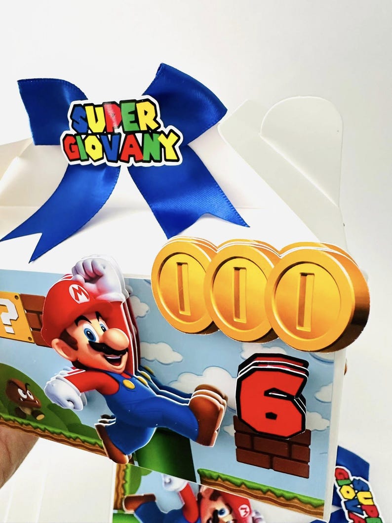 Op de afbeelding: Een witte geschenkdoos versierd met een blauw lint en een kleurrijk "Super Giovany" embleem. De doos heeft een cartoon illustratie van Mario, gouden munten en het nummer 6, allemaal in een levendige, speelse stijl.