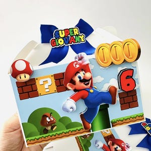Op de afbeelding: Een versierde cadeauverpakking met een Super Mario-thema. De doos heeft een blauwe strik met de tekst "SUPER GIOVANY". De doos is versierd met Mario, munten, een paddenstoel en het cijfer 6. De achtergrond is een blauwe lucht met groene heuvels.