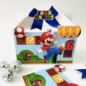 Op de afbeelding: Een witte geschenkdoos met een blauw lint en de tekst "SUPER GIOVANY". De doos heeft een cartoonillustratie van Mario die springt, samen met munten, paddenstoelen en het nummer 6. Extra decoratieve elementen zijn discoballen en een kaart met een gamethema.