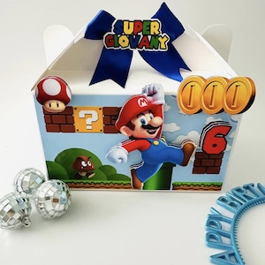 Op de afbeelding: Een witte geschenkdoos versierd met een Super Mario-thema. De doos heeft een blauwe strik, Mario-afbeeldingen, munten en het nummer 6. Extra decoraties zijn discoballen en een blauwe "Happy Birthday" hoofdband.