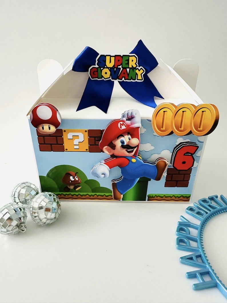 Op de afbeelding: Een witte geschenkdoos versierd met een Super Mario-thema. De doos heeft een blauwe strik, Mario-afbeeldingen, munten en het nummer 6. Extra decoraties zijn discoballen en een blauwe "Happy Birthday" hoofdband.