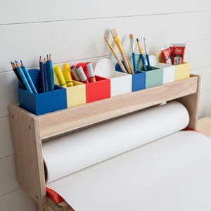 Könnte beinhalten: Künstlerstation aus Holz mit einer Rolle weißem Papier und farbigen Aufbewahrungsfächern für Kunstbedarf. Die Fächer sind blau, gelb, rot, weiß, blau, grün und gelb. Pinsel, Bleistifte und Marker sind sichtbar.