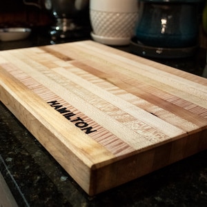 May include: A rectangular wooden cutting board with the word "HAMILTON" printed in black. The board is made of light and dark wood strips. The cutting board is approximately 16 inches long and 12 inches wide.