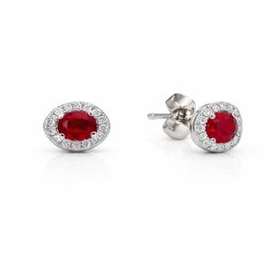 May include: A pair of stud earrings featuring oval-cut, deep red gemstones surrounded by a halo of small, clear gemstones. The earrings are set in a silver-colored metal and are displayed against a white background.