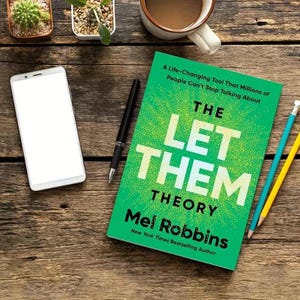 May include: A hardcover book titled "THE LET THEM THEORY" by Mel Robbins, with a green cover and white text, lies on a wooden surface next to a smartphone, pen, pencils, and a cup of coffee. The book's cover also includes the text "A Life-Changing Tool That Millions of People Can't Stop Talking About."