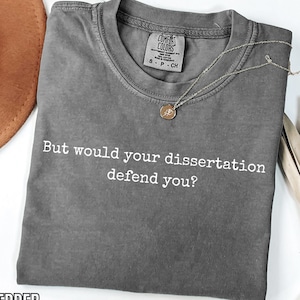 May include: A grey Comfort Colors t-shirt with the text "But would your dissertation defend you?" in white. A gold necklace with a pendant is draped across the shirt. The word "PEPPER" is at the bottom.