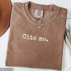 Puede incluir: Camiseta color espresso con el texto blanco "Cite me.". La camiseta tiene cuello redondo y mangas cortas. Un collar plateado con un colgante está drapeado sobre la camiseta. Un sombrero marrón es visible en la esquina superior izquierda.