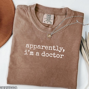 May include: Espresso-coloured t-shirt with the text "apparently, I'm a doctor" in white. A silver necklace with a small pendant rests on the shirt. The shirt is folded on a white surface.