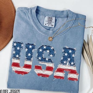 Könnte beinhalten: Ein kurzärmeliges T-Shirt in Jeansblau mit Rundhalsausschnitt und einem Comfort Colors Markenetikett. Auf der Vorderseite befinden sich die Buchstaben USA als Applikation aus ausgefranstem Stoff mit rot-weiß-blauem Flaggenmuster.