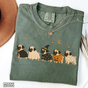 Könnte beinhalten: Ein moosgrünes Kurzarm-T-Shirt mit einer Grafik von fünf Möpsen in Halloween-Kostümen, darunter ein Vampir, ein Geist, eine Hexe, ein Kürbis und eine Mumie. Das T-Shirt hat ein Comfort Colors-Etikett und eine dünne goldene Halskette liegt darauf.