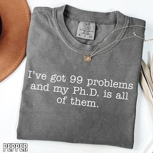 Op de afbeelding: Een grijs Comfort Colors T-shirt met de tekst "I've got 99 problems and my Ph.D. is all of them." Een gouden ketting met hanger ligt op het shirt. Het shirt is opgevouwen en heeft een label "S-P-CH".