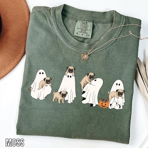 May include: A moss green short-sleeve t-shirt featuring a graphic print of four pugs interacting with white ghost costumes. One pug stands, while others are partially covered by or peeking out from the ghosts. A small orange jack-o'-lantern is included.